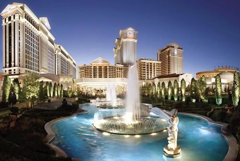 A scenic view of luxury hotel Caesar Entertainment and casino complex featuring a large fountain and illuminated water features surrounded by manicured landscaping during twilight.