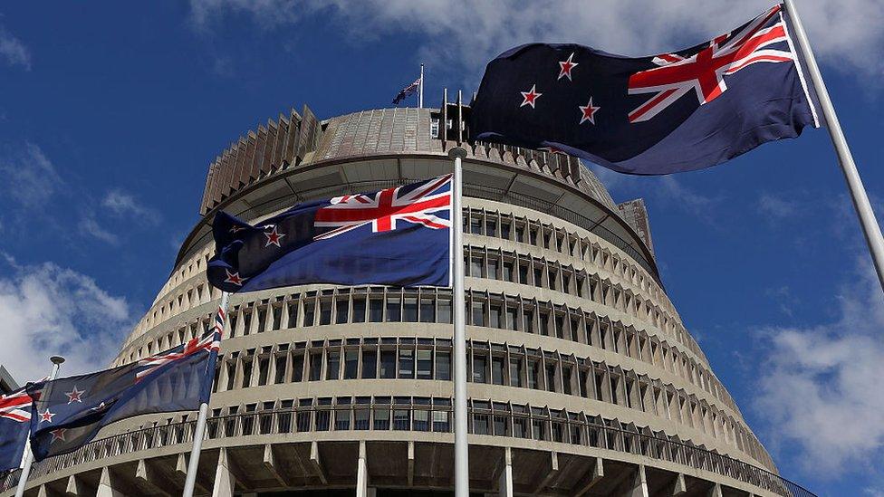 View of the New Zealand Parliament buildings with flags flying in front against a blue sky.