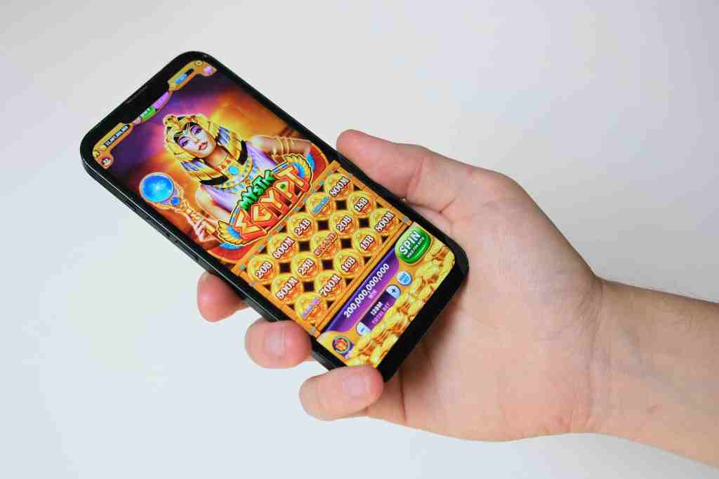 A hand holding a smartphone displaying a colourful slot game featuring an Egyptian theme with various coins and a character.