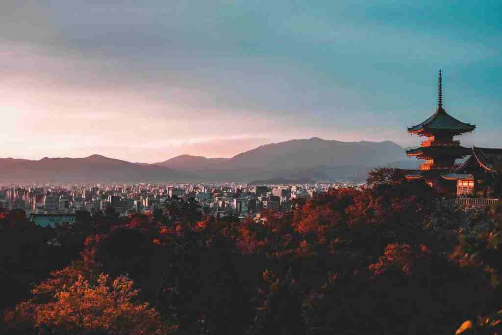 A scenic view of a city with mountains in the background during sunset, featuring a traditional Japanese pagoda in the foreground surrounded by autumn foliage.