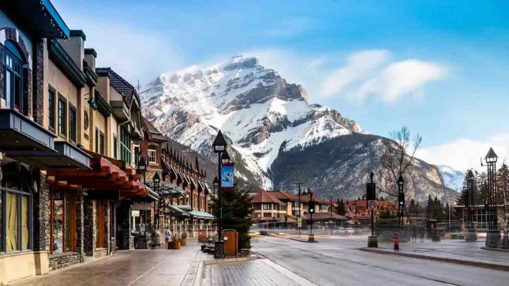 Street view of a charming town with shops and restaurants, framed by snow-capped mountains in the background under a clear blue sky.