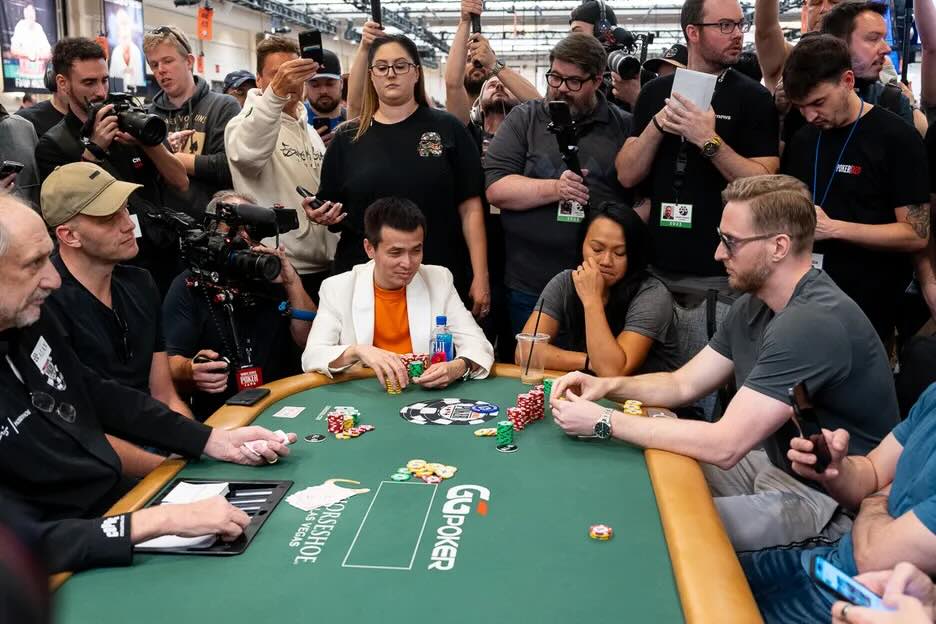 A crowded poker scene with several players at a green poker table, surrounded by a large group of spectators and cameras. The focus is on two players in the foreground, one wearing an orange shirt and white jacket, and the other in a grey shirt, as they engage in a game of Texas Hold'em.